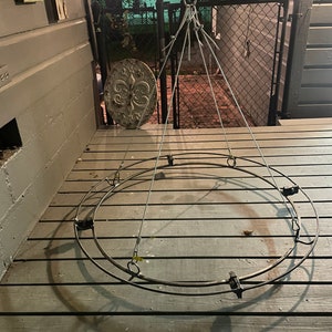 May include: A metal outdoor chandelier frame with a circular design. The frame is suspended by wires from a central point. The frame is resting on a wooden deck. A decorative metal wall hanging with a fleur-de-lis design is visible in the background.