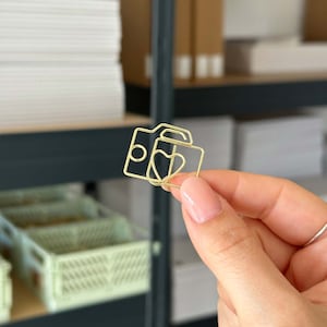 Könnte beinhalten: Ein goldfarbener Papierclip aus Metall in Form einer Kamera mit einem Herzen darin. Der Clip wird von einer Hand gehalten.