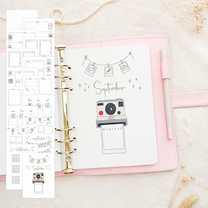 Op de afbeelding: Een roze planner met gouden ringen, open op een pagina met een "September"-ontwerp en een camera-illustratie. Losbladige pagina's met verschillende zwart-witte planningssjablonen zijn links zichtbaar. Een pen rust in de houder van de planner.