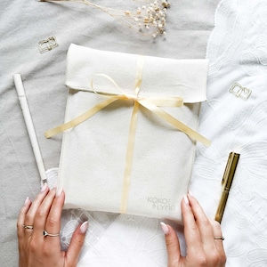May include: A cream canvas pouch tied with a pale yellow ribbon. The pouch is printed with the text "KOKO & LYNN". A gold pen and paperclip are next to the pouch. The pouch rests on a white and grey surface.