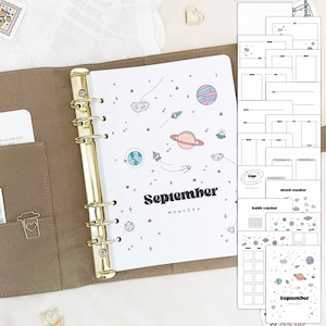 May include: A brown leather planner with gold rings and a white page with a space theme. The page has the word "September" written in black and features planets, stars, and rockets. The planner also includes several blank pages with different layouts for tracking mood, habits, and goals.