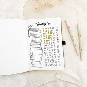 May include: An open notebook with a reading log layout. The page features a stack of illustrated books, titles, star ratings, and heart symbols. Two pens with pink and blue accents are on the right. The words "Reading log" are at the top.