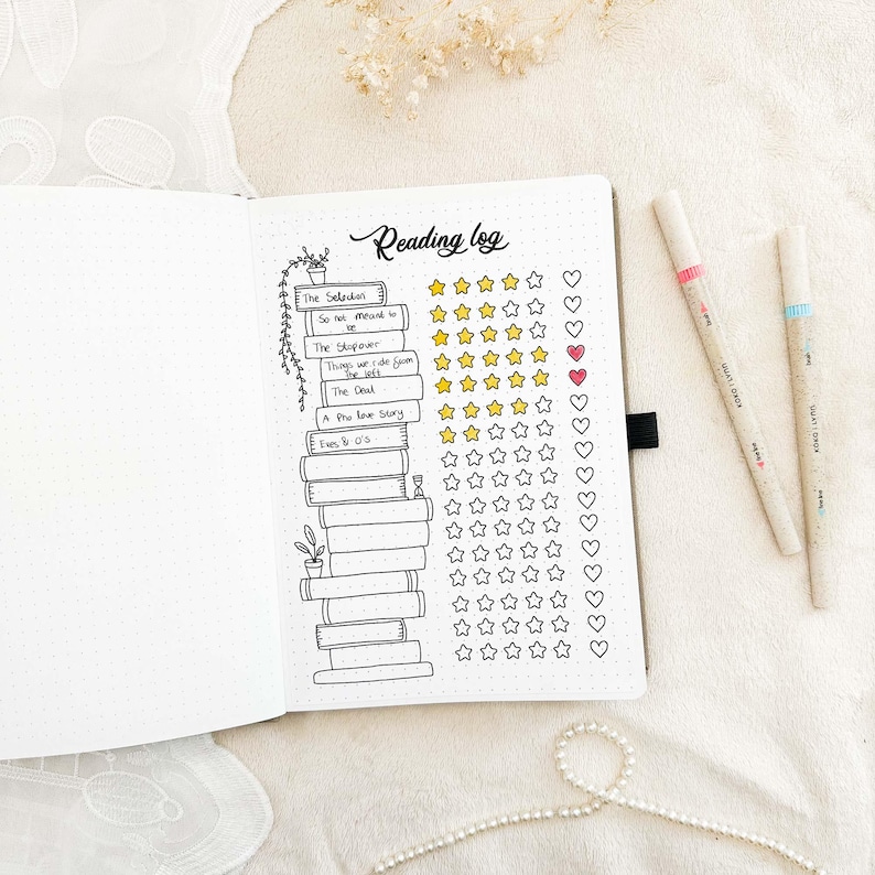 May include: An open notebook with a reading log layout. The page features a stack of illustrated books, titles, star ratings, and heart symbols. Two pens with pink and blue accents are on the right. The words "Reading log" are at the top.