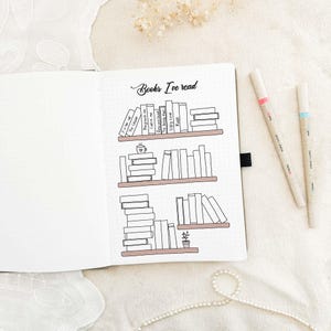 Può includere: Quaderno aperto con un'illustrazione di libreria disegnata a mano e la scritta "Books I've read". L'illustrazione presenta pile di libri sugli scaffali, una tazza di caffè e una piccola pianta. Due penne con dettagli rosa e blu sono a destra.