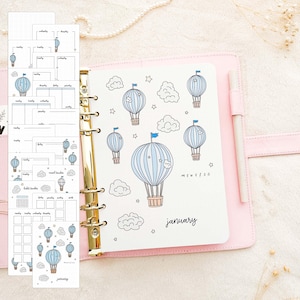 Op de afbeelding: Een roze planner met gouden ringen, open op een pagina met blauwe luchtballon illustraties en het woord "januari". Extra plannerpagina's met kalenderindelingen en ballonontwerpen zijn zichtbaar. Een witte pen rust in de planner.