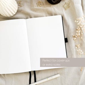 May include: Open, blank notebook with dot grid pages. The notebook is black with a black ribbon bookmark. The text "Perfect for cover-ups or extra notes" is visible. Decorative gold paper clips and a pen are also in the image.