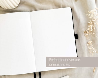 May include: Open, blank notebook with dot grid pages. The notebook is black with a black ribbon bookmark. The text "Perfect for cover-ups or extra notes" is visible. Decorative gold paper clips and a pen are also in the image.