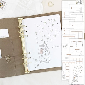 Op de afbeelding: Een bruine lederen planner met gouden ringen. De planner heeft een transparante pagina met een zwart-witte illustratie van een pot vol met vuurvliegjes. De pagina bevat ook de tekst "M T W T F S" en "Juli".