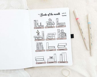 May include: An open notebook with a hand-drawn "Books of the month" layout. The layout features illustrated bookshelves for each month, with book titles and decorative elements. Two pens with pink and blue accents are on the right.