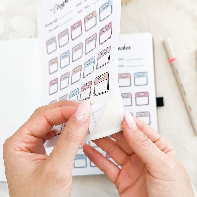 May include: A sheet of stickers with scale illustrations in pink, blue, and peach hues. The stickers are being peeled from a notebook page. A pen is visible in the background, suggesting a planner or journal theme.