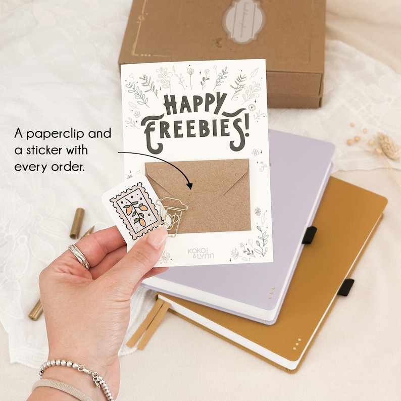 May include: A white card with the text "Happy Freebies!" in black lettering. A brown envelope is attached to the card with a silver paperclip. A sticker with a drawing of three lemons on a branch is attached to the envelope. The card is on top of a purple journal and a yellow journal.