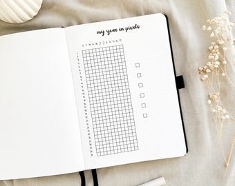 May include: A black and white planner page with the title "my year in pixels". The page is divided into a grid with 31 rows and 12 columns, representing the days of the month and the months of the year. There are also 5 empty boxes on the right side of the page.