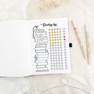 May include: Open notebook with a reading log layout. The title "Reading log" is handwritten at the top. The log includes book titles, star ratings, and heart symbols. Two pens with pink and blue accents are on the right.
