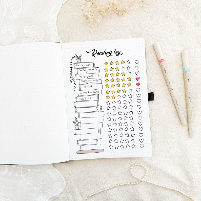 May include: Open notebook with a reading log layout. The title "Reading log" is handwritten at the top. The log includes book titles, star ratings, and heart symbols. Two pens with pink and blue accents are on the right.