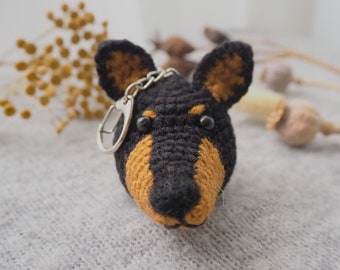 Op de afbeelding: Een gehaakte sleutelhanger in de vorm van een zwarte en bruine hond met een zilveren sleutelring. De hond heeft zwarte ogen en een bruine neus.