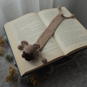 May include: A crocheted dog bookmark in brown and beige with a black nose and brown eyes. The bookmark is placed in an open book.