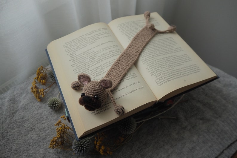 May include: A crocheted dog bookmark in brown and beige with a black nose and brown eyes. The bookmark is placed in an open book.