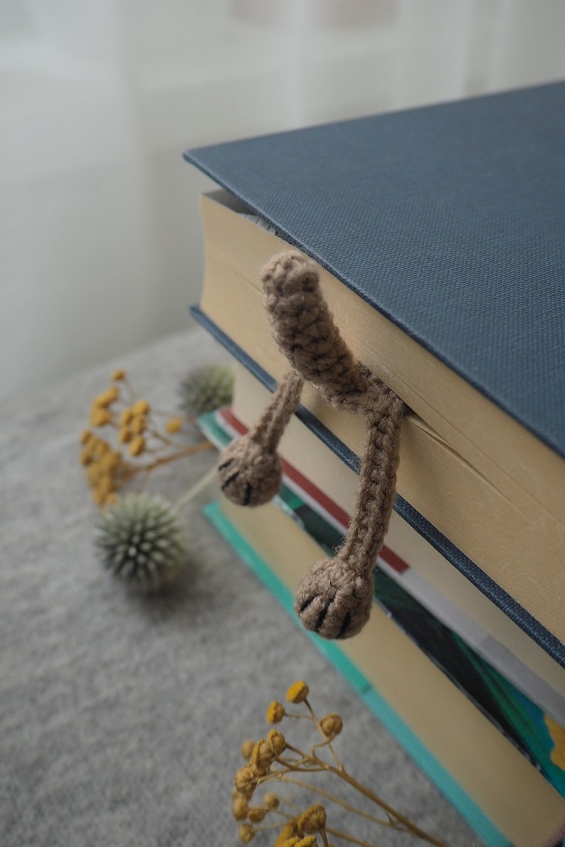 May include: A brown crocheted bookmark shaped like a cat's paw with two paws sticking out from the top of a stack of books. The books are on a gray surface with dried flowers.