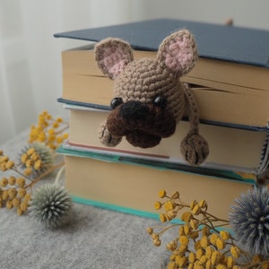 May include: A crocheted brown and tan dog bookmark peeking out from a stack of books. The books are on a gray surface with yellow flowers.