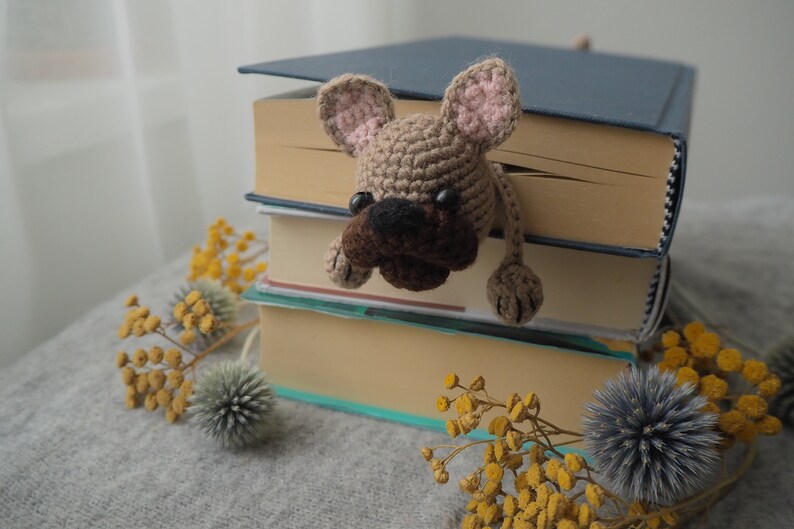 May include: A crocheted brown and tan dog bookmark peeking out from a stack of books. The books are on a gray surface with yellow flowers.
