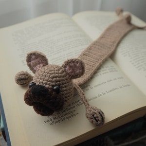 May include: A crocheted dog bookmark in brown and pink with black eyes. The bookmark is laying on a book with text in French.