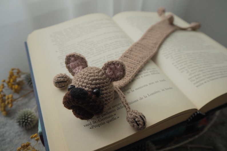 May include: A crocheted dog bookmark in brown and pink with black eyes. The bookmark is laying on a book with text in French.