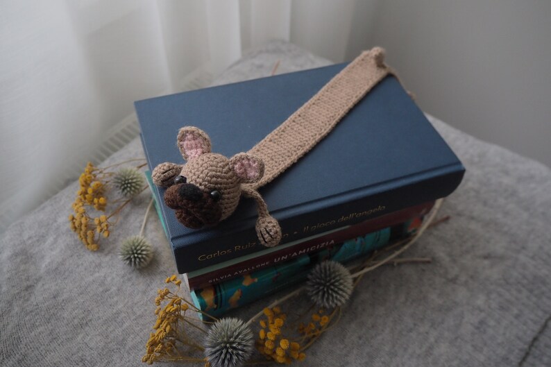 May include: A crocheted brown and pink dog bookmark with a long tail, sitting on top of a stack of books. The top book is blue and has the text "Carlos Ruiz Zafon" on the spine.
