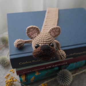 May include: A crocheted brown and beige dog bookmark with black eyes and a brown nose. The bookmark is on top of a stack of books.
