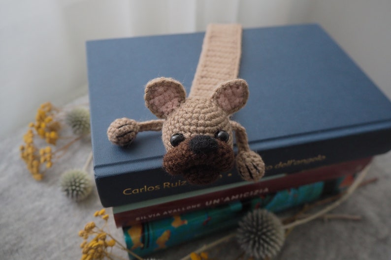 May include: A crocheted brown and beige dog bookmark with black eyes and a brown nose. The bookmark is on top of a stack of books.