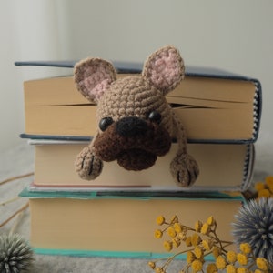 May include: A crocheted brown and pink French bulldog bookmark peeking out from between three books. The books are stacked on top of each other and have brown covers.