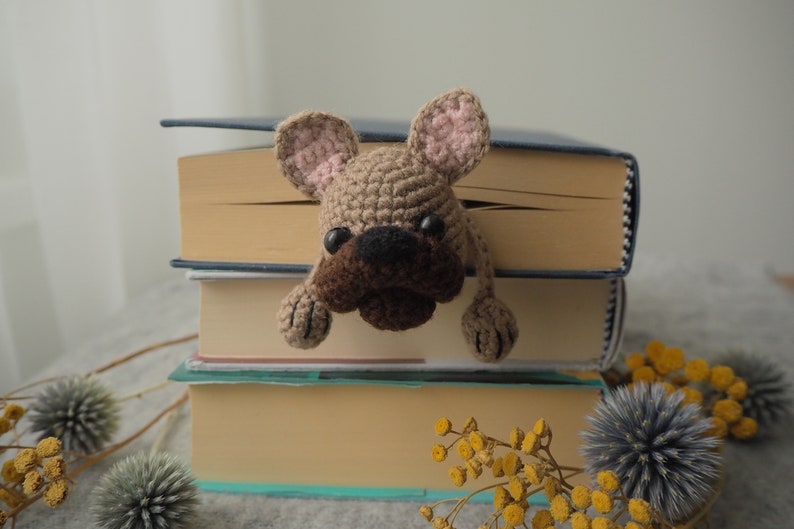 May include: A crocheted brown and pink French bulldog bookmark peeking out from between three books. The books are stacked on top of each other and have brown covers.