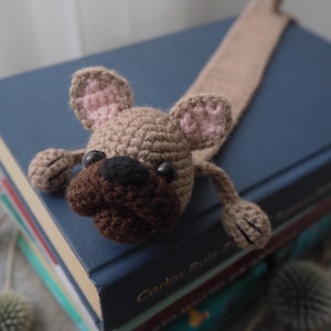 May include: A crocheted dog bookmark with brown fur, a black nose, and pink ears. The bookmark is laying on a stack of books.