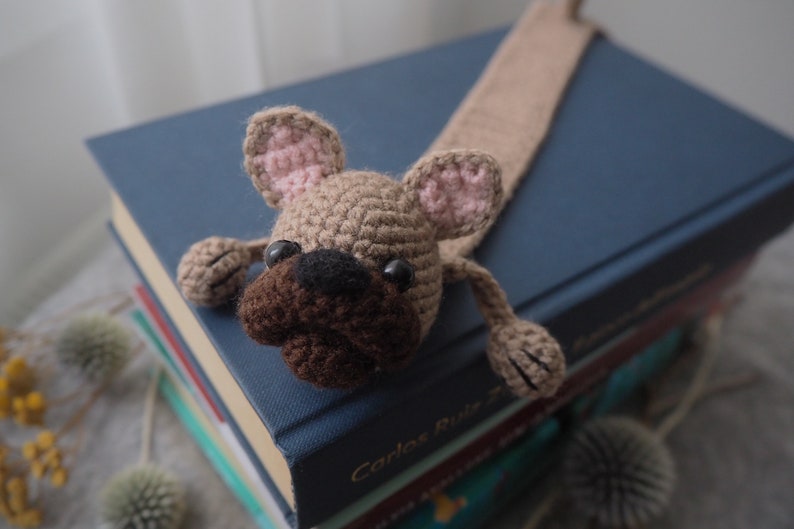May include: A crocheted dog bookmark with brown fur, a black nose, and pink ears. The bookmark is laying on a stack of books.