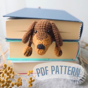 Könnte beinhalten: Ein gehäkelter braun-beiger Dackel, der über einen Stapel Bücher schaut. Der Hund hat schwarze Augen und eine schwarze Nase. Der Text "PDF PATTERN" befindet sich in einer weißen Blase unten rechts.