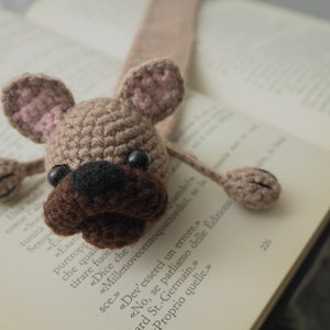 May include: A crocheted brown and pink dog bookmark with black eyes. The bookmark is in the shape of a dog with floppy ears and a long tail.