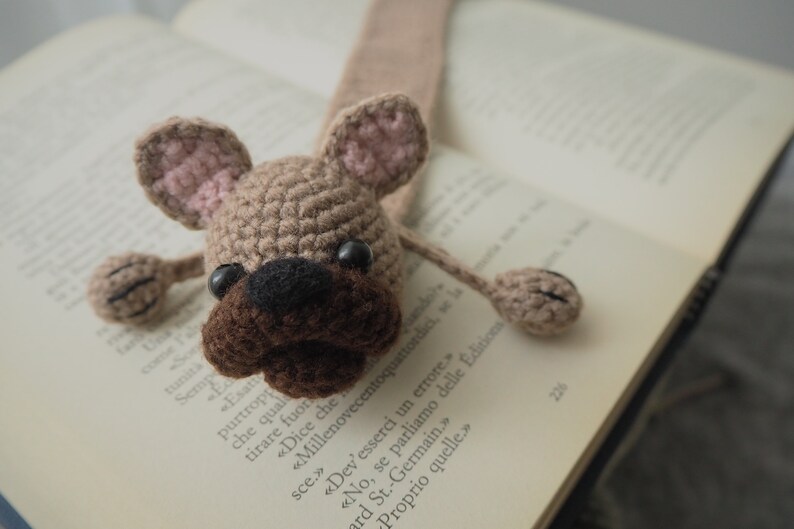 May include: A crocheted brown and pink dog bookmark with black eyes. The bookmark is in the shape of a dog with floppy ears and a long tail.