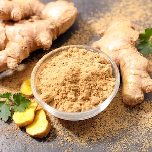 May include: A clear glass bowl filled with light brown ginger powder, surrounded by fresh, knobby ginger roots, sliced ginger pieces, and small green herb leaves, with loose ginger powder scattered across the dark gray surface.