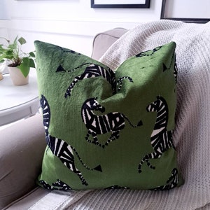 May include: A square green decorative pillow with a black and white zebra pattern. The pillow is placed on a gray sofa with a white knitted throw blanket. Decorative artwork hangs on the wall in the background.