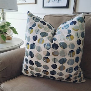 May include: A throw pillow with a white background and a pattern of blue, green, and gold circles. The pillow is sitting on a brown couch.