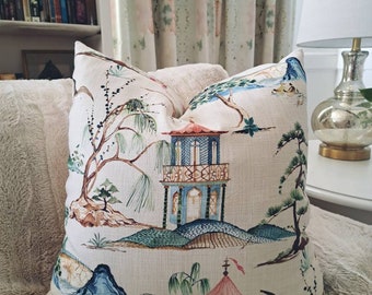 May include: A decorative throw pillow with a beige background and a colorful Asian-inspired design. The pillow features a blue and green pagoda, a red and pink tent, and a variety of trees and mountains.