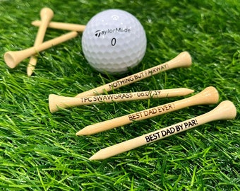 Può includere: Una pallina da golf con il numero 0 e il marchio TaylorMade, accanto a quattro tee da golf in legno. I tee riportano le scritte "NOTHING BUT FAIRWAY!", "TPC SWAWGRASS - 06.07.19", "BEST DAD EVER!" e "BEST DAD BY PAR!"