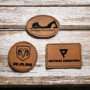 Peut inclure: Trois patchs en cuir marron avec du texte noir et des images. Le patch du haut a une silhouette d'excavatrice et le texte "Pettipas Excavation". Le patch de gauche a un logo de tête de bélier et le texte "RAM". Le patch de droite a une lettre "P" stylisée et le texte "Pettipas Engraving".
