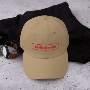 May include: A tan baseball cap with a red embroidered rectangle that says "No Entiendo" on it.