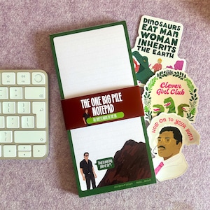 May include: A notepad with a green border and the text "THE ONE BIG PILE NOTEPAD" on a red band. The notepad has lined pages and is accompanied by stickers with dinosaur and quote designs. A keyboard is visible in the background.