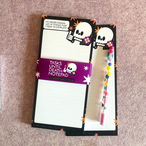 May include: A notepad with a skull design and the text "TASKS UNTIL DEATH NOTEPAD". The notepad has lined pages and a purple band. A pen with colorful polka dots is also visible.