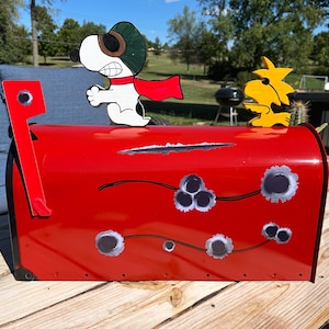 May include: A red metal mailbox with Snoopy and Woodstock figures on top. The mailbox has bullet hole designs and scratch marks. Snoopy is wearing a pilot's hat and red scarf. Woodstock is yellow.