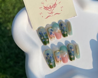 May include: Set of ten false nails with a green and pink ombre design. Each nail features a gold accent and a hand-painted floral design with blue flowers.