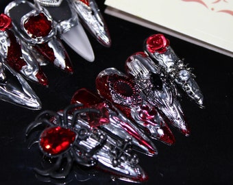 May include: Close-up of a set of fake nails with a gothic theme. The nails are decorated with silver, red, and black accents, including a spider, roses, and hearts.