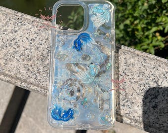 May include: A clear phone case featuring a decorative design of blue and light blue goldfish swimming amidst shimmering, iridescent accents and small butterfly shapes. The case has a rectangular cutout for a camera lens at the top.