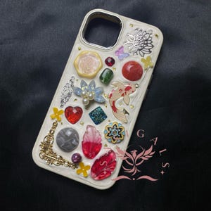 May include: White iPhone case decorated with colorful embellishments, including a red heart, a gold crown, a blue flower, a red and white leaf, a gold fish, and a gold key.